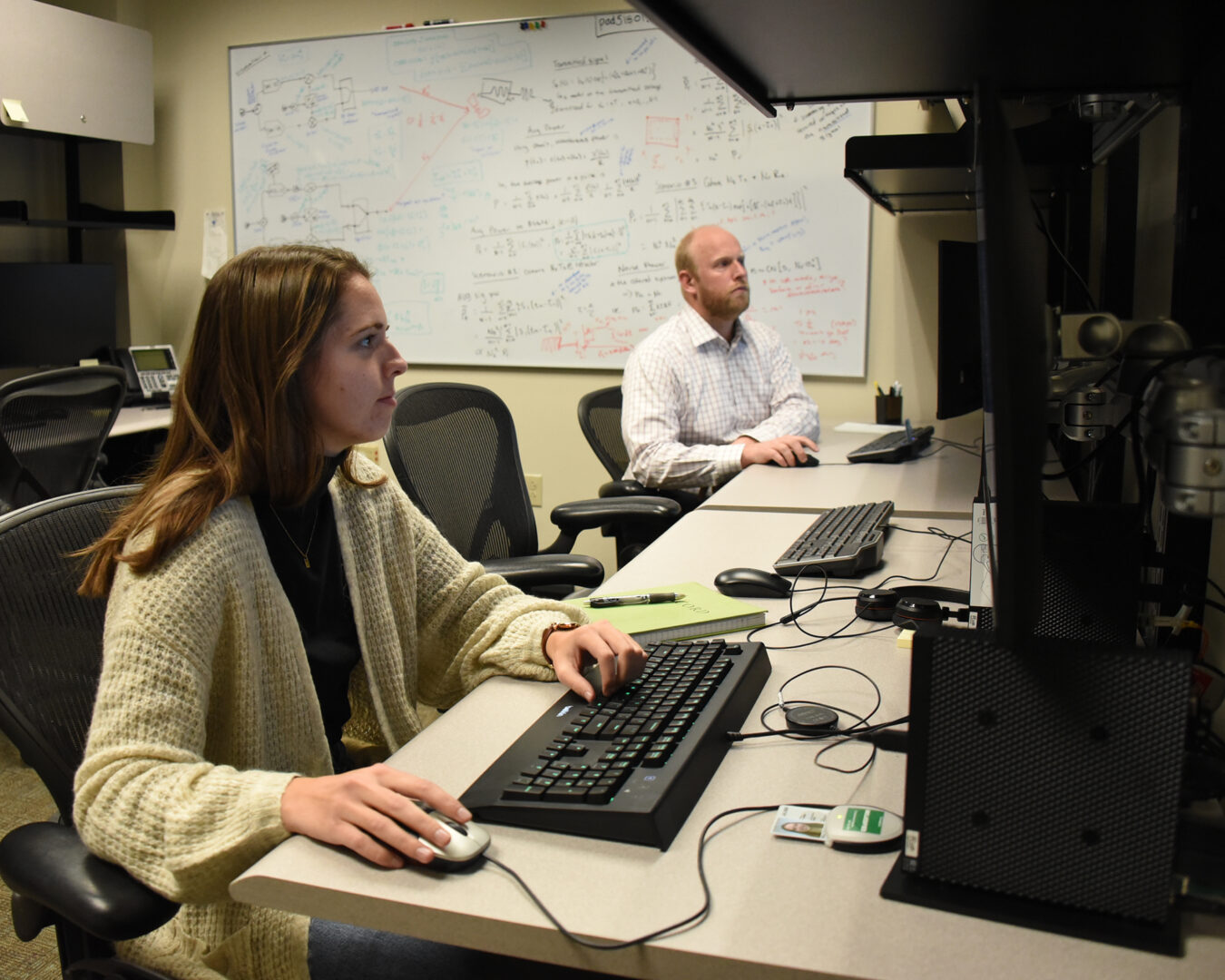 two people work on computers. A formula-filled whiteboard is on the wall behind them.